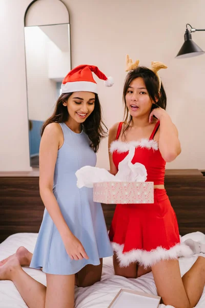 Two young Asian girls (one Indian, one Chinese) exchange Christmas gifts. One is wearing a red Santarina outfit and the other a blue dress with a Santa hat. They are both smiling happily.
