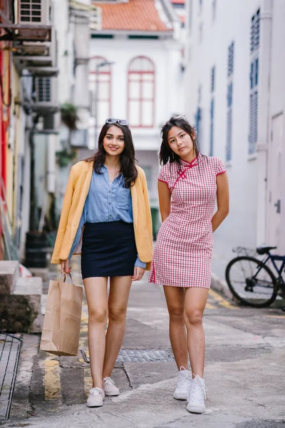 two close friends walking together down an alley. They are both wearing trendy retro clothing and holding hands as they walk happily together. One is Chinese, the other Indian