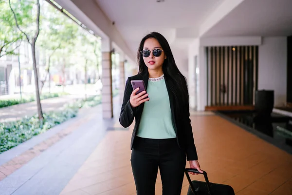 Portrait of an elegant and beautiful young Asian woman waiting on the side of the street for her ride that she booked via a ride-hailing app. She is holding onto a luggage suitcase on wheels.