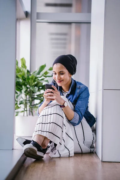 Genç bir Ortadoğulu Müslüman kadın gün boyunca bir ofiste, ortak çalışma alanında veya kütüphanede oturuyor ve akıllı telefonunu kullanıyor. Başörtüsü takıyor (başörtüsü)).