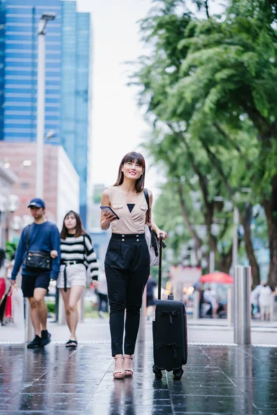 迷人的亚洲华裔女性在智能手机上通过她的骑行欢呼应用预订了一本书 她拿着行李走着 穿着工作服 穿着时髦的衣服 — 图库照片