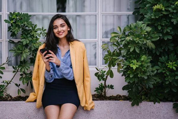 时尚的印度亚洲女性在智能手机上发短信 白天她靠在窗台上 她穿着复古的黄色夹克而不是休闲服 — 图库照片