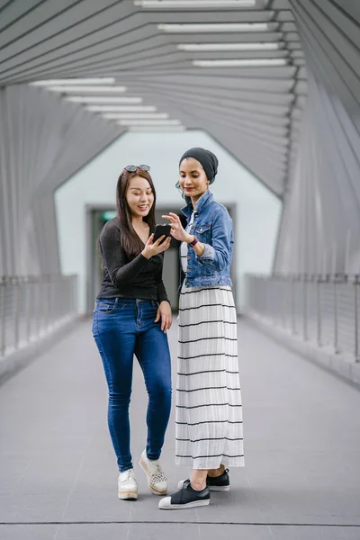 白天两个不同种族的朋友在一座桥上 一个是马来族穆斯林妇女 另一个是亚裔中国人 他们都很年轻 精力充沛 很有魅力 — 图库照片