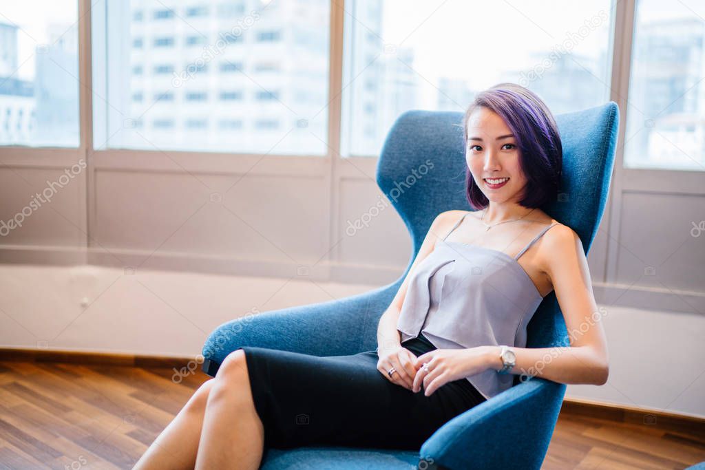 A young, slim and elegant Asian Chinese woman in a professional dress and dyed purple hair sits in a red armchair in her office coworking space. She is smiling candidly.