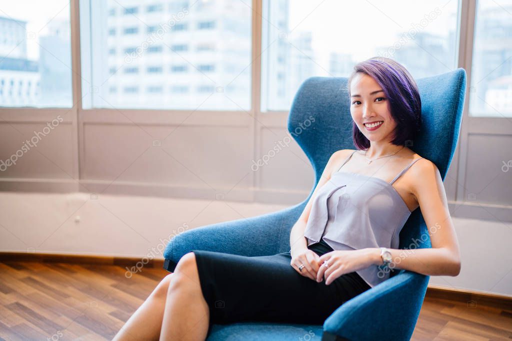 A young, slim and elegant Asian Chinese woman in a professional dress and dyed purple hair sits in a red armchair in her office coworking space. She is smiling candidly.