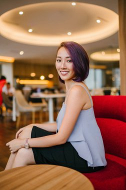 Profesyonel elbiseli ve boyalı mor saçlı genç, ince ve zarif bir Asyalı kadın çalışma odasında kırmızı bir koltukta oturuyor. İçtenlikle gülümsüyor..