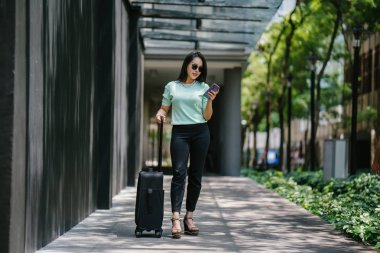 O bir yolculuk dolu uygulama üzerinden rezervasyonu onu binmek için caddenin kenarında bekleyen zarif ve güzel bir genç Asyalı kadının portresi. Tekerlekli bir bavula tutunuyor.. 