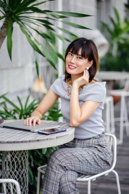 Açık havada şık bir yerde oturan bir emin, şık ve çekici orta yaşlı Çinli Asyalı kadın portresi. O bir pohpohlama gri kıyafeti giyiyor.