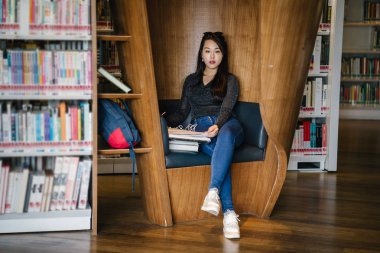 Genç, güzel ve çekici Çinli Asyalı değişim öğrencisi kütüphanedeki kahverengi bir kulübede okuyor ve çalışıyor..