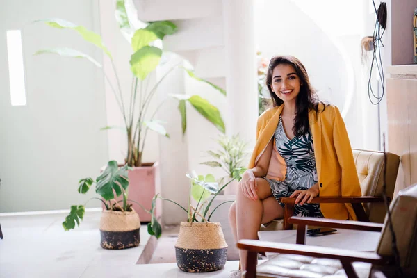  şık, güzel ve genç Hintli Asyalı kadın elbise ve omuzları üzerinde bol dökümlü bir sarı ceket. O şık iç mekanlara bir sandalyede oturuyor