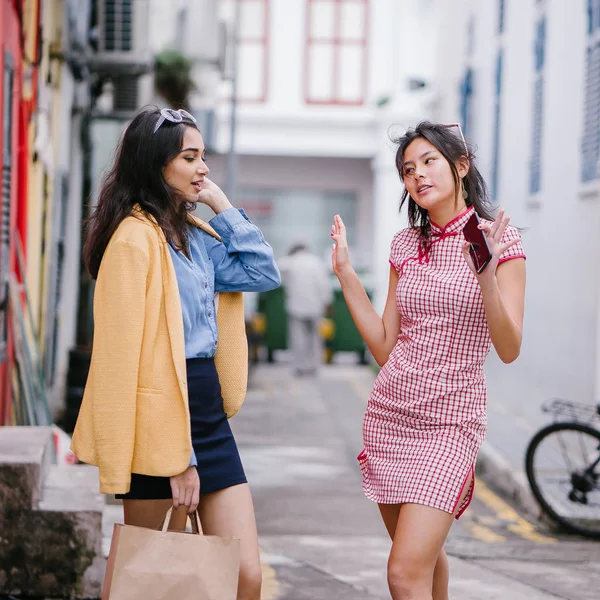 two close friends walking together down an alley. They are both wearing trendy retro clothing and holding hands as they walk happily together. One is Chinese, the other Indian