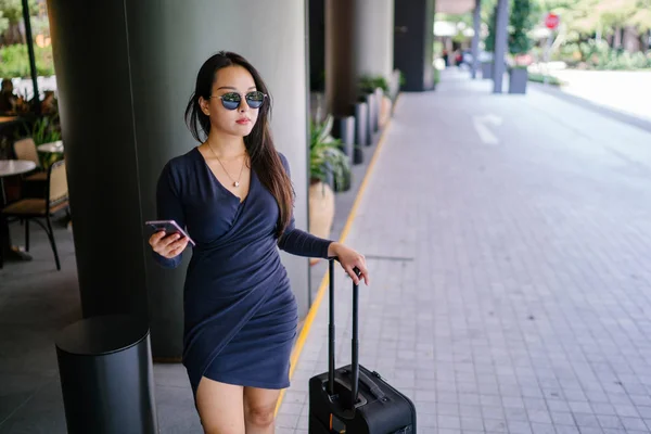 Portrait of an elegant and beautiful young Asian woman waiting on the side of the street for her ride that she booked via a ride-hailing app. She is holding onto a luggage suitcase on wheels.