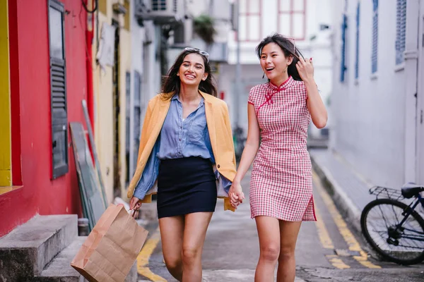 two close friends walking together down an alley. They are both wearing trendy retro clothing and holding hands as they walk happily together. One is Chinese, the other Indian