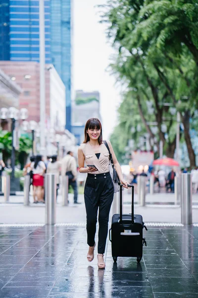 attractive Asian Chinese woman books a ride through her ride hailing app on her smartphone. She is walking with her luggage and is stylishly dressed in work wear