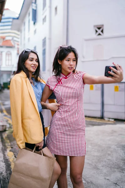 iki genç Asyalı kız bir selfie birlikte gündüz sokakta almak. Onlar çeşitli çifti bir arkadaş, bir Çince bir qipao olarak ve diğer Hint retro bir kıyafeti vardır. 