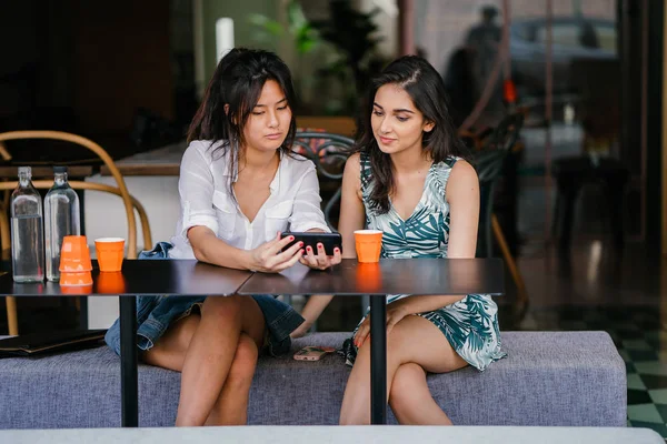 Two close friends (women) sit in a cafe during the day and watch a video together on a smartphone. They are both smiling as they enjoy the video and each other\'s company.