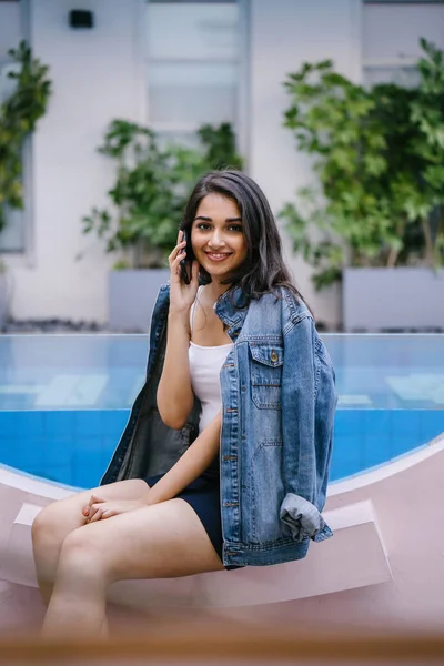 Akıllı telefonda konuşurken bir tatil havuzu tarafından gün boyunca oturan genç bir Hint Asyalı kadının bir yaşam tarzı portre