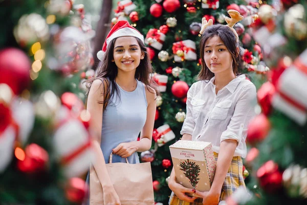 Two close friends exchange Christmas presents with one another on the holiday season. They are both Asian, one is Indian, the other Chinese. They are smiling happily as they exchange gifts.