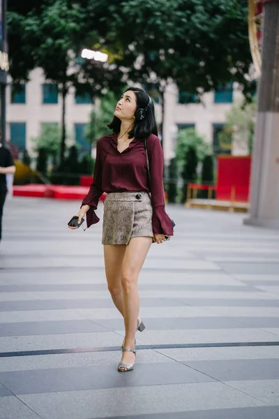 beautiful Korean woman in an elegant blouse and shorts dancing as she walks down a street in the city with her headphones on