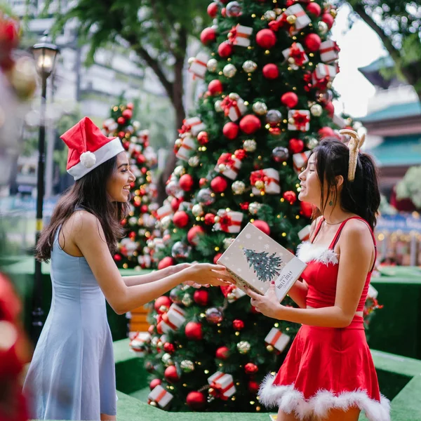 Two young Asian girls exchange presents with one another under a decorated Christmas tree in a street in the city during the day. One is Indian, the other Chinese