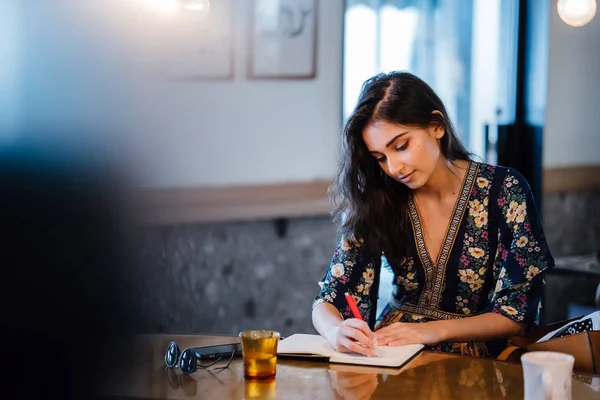 Genç Hintli Asyalı kadın bir kafede oturur ve yazıyor üstünde onun defter onun kalemi ile