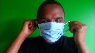 Mavi cerrahi maske takan bir adam. Afrikalı. Sade. Ön manzara, kapatın. Kayışlar kulakların üzerinde. Yüz maskesi ayarlanıyor. Kameraya bakıyorum. Koronavirüs için koruyucu teçhizat, covid-19, 2019-ncov. Yeşil arka plan, yeşil ekran konsepti. Gölge. Krom anahtar.