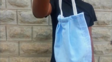 Genç, yetişkin bir adam mavi bir maske takıyor. Yakın çekim. Afrika kökenli Amerikalı bir adam tek eliyle kamera karşısında maske takıyor. Vermek için elini uzat. Etnik köken. Çeşitlilik. Covid-19 konsepti.