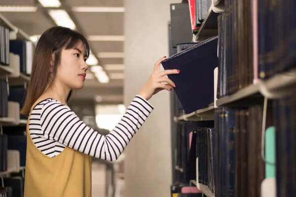 smart woman college student searching books; portrait of focused woman university student studying, researching, reading, finding book or textbook in library; asian young adult woman model
