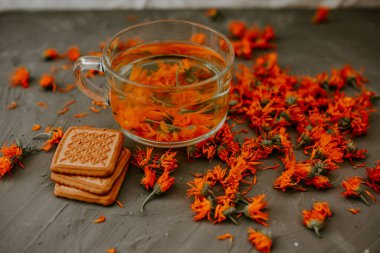 Calendula çiçekli ve bisküvili çay. Şeffaf cam bardak ve çay tabağı. Şifalı bitkisel kurutulmuş bitkiler kadife çiçeği, turuncu takvim. Tarafsız beyaz ve gri arkaplan.