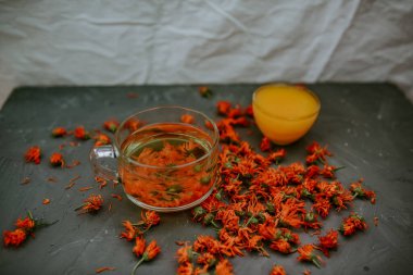 Büyük şeffaf bir kupada çiçek çayı. Küçük bir fincan ağzına kadar sarı sıvı bal ile doldurulur. Şifalı bitkisel kurutulmuş bitkiler kadife çiçeği, turuncu takvim. Tarafsız beyaz ve gri arkaplan.
