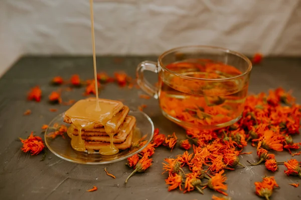 The honey flows in a thin stream onto the baked goods. Orange calendula flower tea in a transparent cup and honey-covered cookies in a saucer. Medicinal herbal dried plants marigold,.gray background.