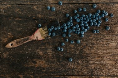 Mavi taze yaban mersini eski kahverengi ahşap çatlamış bir masaya saçılmış. Mavi boyalı bir boya fırçası meyvenin yanında yatıyor..