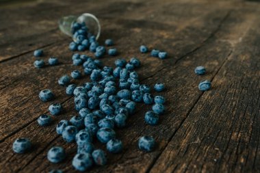 Taze mavi mat yuvarlak yaban mersini şeffaf bir bardakla doldurulmuş, ahşap kahverengi eski püskü bir masa üzerinde duruyor ve böğürtlenler her yere saçılmış.