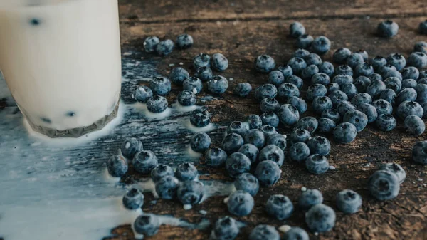 Berries are thrown into the transparent glass making splashes of milk. Blue fresh blueberries are scattered on an old brown wooden cracked table. White water pour down on the table.