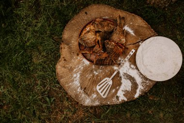 Kızarmış balık haçbalığı sazanı, üzerine beyaz un serpilmiş ve bir mutfak spatulası serpiştirilmiş eski bir ahşap kütüğün üzerinde bir tabakta yatıyor. Yeşil çimenler. Yaz. piknik.