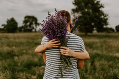 Çizgili tişörtlü esmer bir adam arkası dönük duruyor ve bir kadına sarılıyor. Kız elinde büyük bir demet leylak ve menekşe çiçeği tutuyor. Arka planda yeşil sarı çimenler ve ağaçlar var..