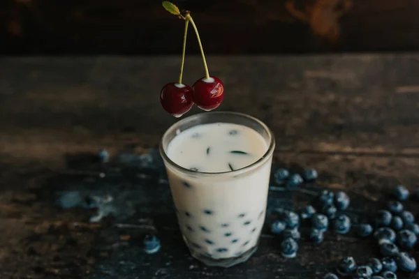 a womans hand holds a cherry and soaks it in milk. blueberries are scattered