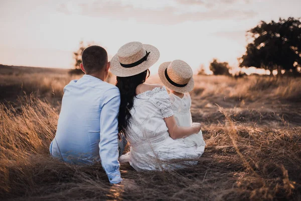 Far, brunett mor och liten dotter sitter på torrt gräs i vita klänningar — Stockfoto