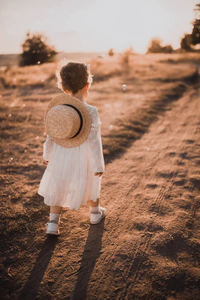 little girl in a white dress with curly hair goes towards the sun at sunset