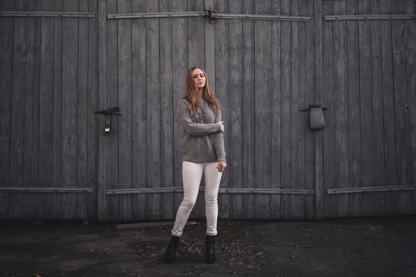 Frau in weißer Hose grauer Strickpullover schwarze Stiefel auf grauem Holztor — Stockfoto