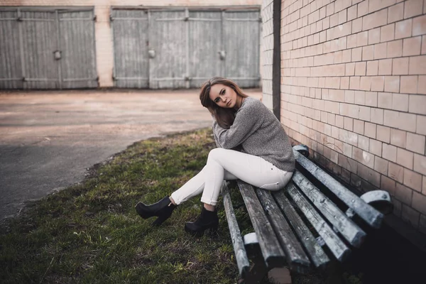 화이트 팬츠를 입은 초라 한 벤치에 앉아 있는 여자 회색 뜨개질 스웨터검은 부츠 — 스톡 사진