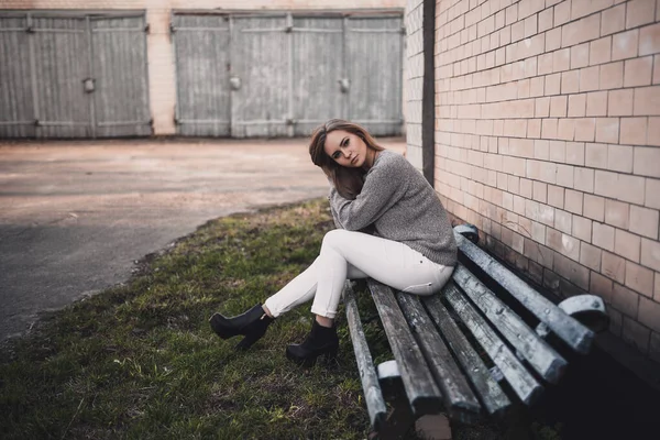 Femme assise sur un banc minable en pantalon blanc.gris tricoté pull bottes noires — Photo