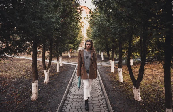 Mujer en pantalones blancos y un abrigo marrón cerca de los árboles — Foto de Stock