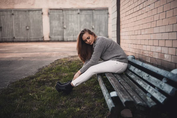 Mujer sentada en un banco de mala calidad en blanco pants.gray suéter de punto botas negras — Foto de Stock