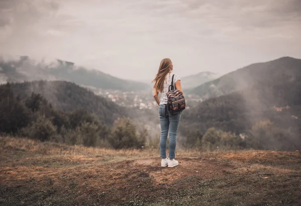 Young blonde woman standing alone with backpack on wild forest mountains