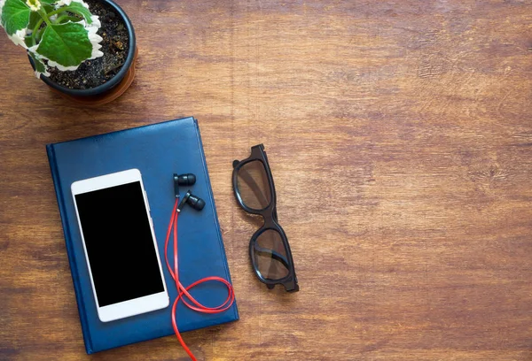 Smartphone, blue notebook, black eyeglasses, red earphones and green flower on wooden table. Top view with copy space, flat lay