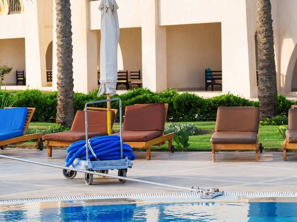 Manual equipment for cleaning pool: brush and hose with brown deckchairs on the background
