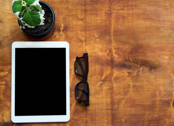 White digital tablet with black glasses and green plant on wood table, top view. Copy space, flat lay