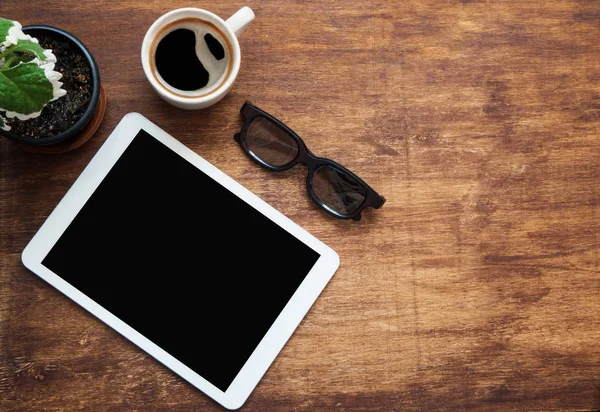 Tablet blank screen, black glasses cup of coffee and green flower on wood desk. Copy space, flat lay