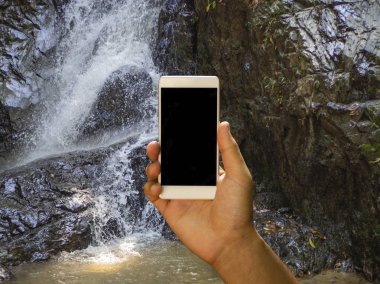 Bir erkek eli boş siyah perde ve arka planda odaklı şelale ile beyaz cep telefonu tutan mockup görüntü